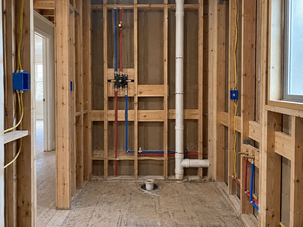 Bathroom at the rough-in stage with open stud walls, new supply lines, a drain and vent stack and a shower valve set between studs